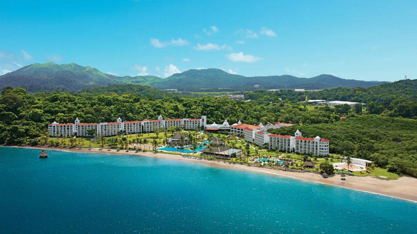 Aerial view of Dreams Playa Bonita Panama on the beach, with pools, palm trees and green mountains behind